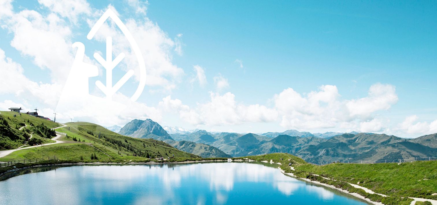 Klarer Bergsee umgeben von grünen Hügeln und Bergen unter blauem Himmel. Links oben sind ein stilisiertes Blatt und KitzSki Gams als Symbole zu sehen.