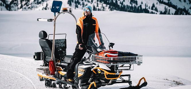 Ein Mann in orange-schwarzer Kleidung steht auf einem Schneemobil vor schneebedeckten Bergen und Bäumen. Der Himmel ist klar und blau.
