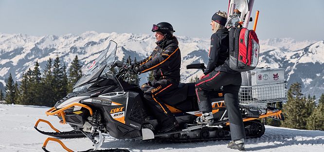 Zwei Personen fahren auf einem Schneemobil durch verschneite Berge. Die vordere Person lenkt, die hintere steht und trägt Ausrüstung. Schnee bedeckte Landschaft.