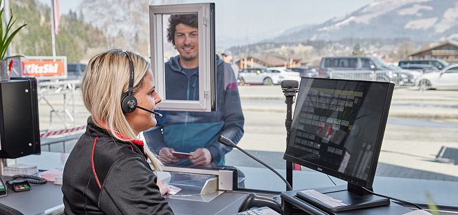 Eine Frau mit Headset sitzt an einem Ticketschalter, während ein Mann am Fenster ein Ticket kauft. Im Hintergrund sind Berge zu sehen.