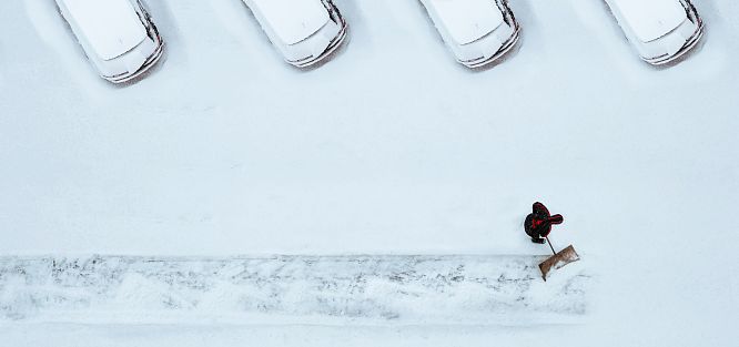 Person schaufelt Schneeweg neben geparkten Autos von oben. Alles ist schneebedeckt und die Szene zeigt eine winterliche Stimmung mit klaren Linien.