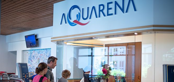 Menschen stehen am Empfang der Aquarena. Großer Schriftzug "AQUARENA" mit Wassermotiv im Hintergrund. Helle, freundliche Atmosphäre mit Glasfenster.