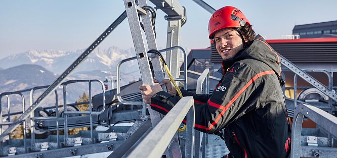 Ein Mann mit rotem Helm und schwarzer Jacke steht in einer Seilbahnstation, schneebedeckte Berge im Hintergrund, sonniges Wetter.